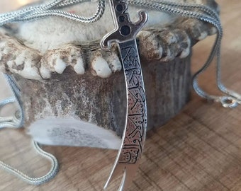 May include: A silver necklace with a sword pendant. The sword is engraved with Arabic calligraphy.