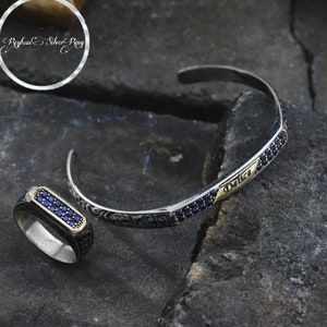 May include: A silver ring and bracelet set with blue gemstones. The ring has a rectangular setting with a row of blue gemstones. The bracelet has a delicate design with a row of blue gemstones and the word "Delice" engraved on it.