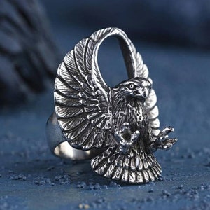 Puede incluir: Un anillo de plata con un diseño de águila detallado. Las alas del águila están extendidas y su cabeza está girada hacia un lado. El anillo está hecho de metal y tiene un acabado pulido.