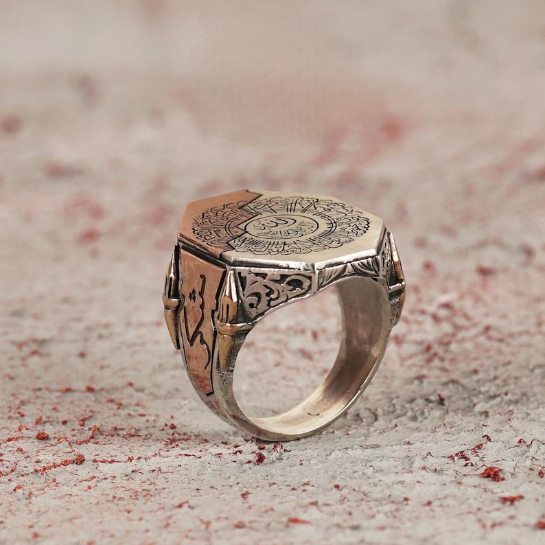 May include: A silver and copper-toned ring featuring an octagonal face with intricate Arabic calligraphy engravings. The band displays detailed scrollwork patterns on the sides, with a contrasting metallic finish that highlights the engraved text.