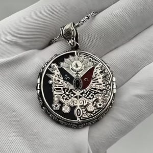 Puede incluir: Un collar con un colgante de plata que presenta un diseño detallado y ornamentado del escudo de armas del Imperio Otomano. El colgante es circular y presenta un fondo de esmalte negro y rojo con intrincados detalles en plata.