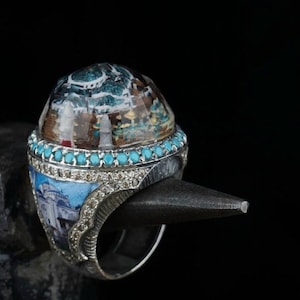 Op de afbeelding: Een zilveren ring met een turquoise en heldere stenen zetting. De steen heeft een gedetailleerd miniatuur schilderij van een gebouw met een koepel.
