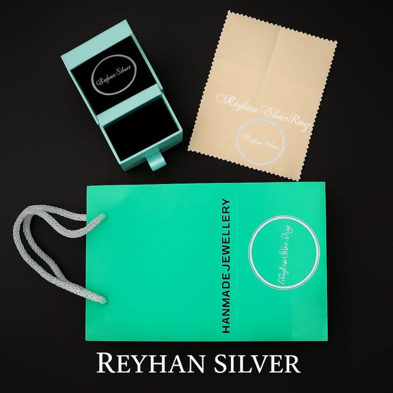 May include: A turquoise jewelry gift bag with white rope handles and the text HANMADE JEWELLERY, a matching turquoise ring box with a black interior, and a beige polishing cloth featuring the text Reyhan Silver Ring and Reyhan Silver.