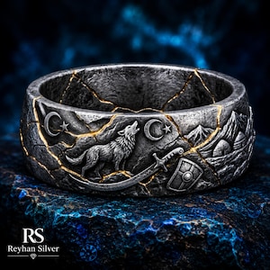 May include: A wide silver ring with a dark, textured finish featuring a howling wolf, a curved sword, a shield, crescent moons, and mountain motifs. The surface is accented with gold-colored cracks throughout the design, creating a segmented look.