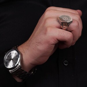 May include: A silver-toned ring featuring an octagonal face with intricate engraved circular patterns and side detailing, worn alongside a silver-toned analog wristwatch with a metallic link band and a white face with black numerals.