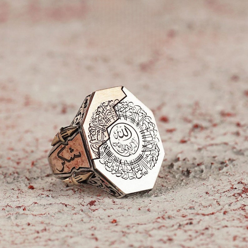 May include: A silver-toned metal ring featuring an octagonal face engraved with intricate Arabic calligraphy. The sides of the ring display additional etched script and decorative metalwork, showcasing a polished finish with dark recessed details.