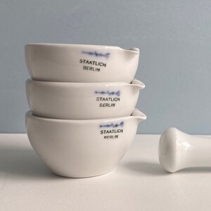 Puede incluir: Un juego de tres morteros y mazos de porcelana blanca, apilados uno encima del otro. Los cuencos están etiquetados como "Staatlich Berlin". El mazo también es de porcelana blanca.