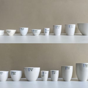 May include: Two rows of white porcelain crucibles of varying sizes. The crucibles are marked with black text and blue lines. The crucibles are arranged in order of size, with the smallest crucibles on the left and the largest crucibles on the right.
