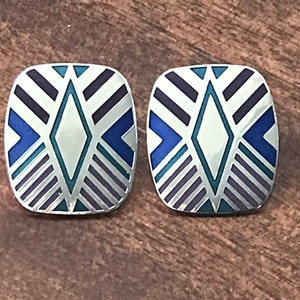 May include: A pair of square silver-toned stud earrings. Each earring features a geometric design with a central diamond shape, surrounded by blue, teal, and purple accents. The earrings are set against a wooden surface.