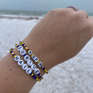 Pode incluir: Três pulseiras de contas com as palavras "U of M", "Go Blue" e "Hail" em letras brancas sobre um fundo azul e amarelo.