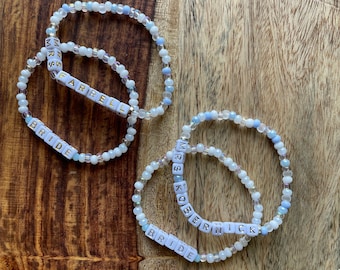 May include: Two beaded bracelets with the words "Bride" and "Mrs. [Name]" spelled out in white beads. The bracelets are on a wooden surface.