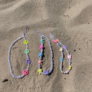 Puede incluir: Tres collares de perlas con abalorios coloridos. El primer collar tiene signos de paz, el segundo tiene osos de dibujos animados y el tercero tiene mariposas.
