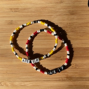 May include: Two beaded bracelets with black, white, red, and yellow beads. The bracelets spell out "AS IF" in black letter beads.