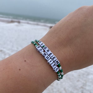 Puede incluir: Dos pulseras de cuentas con cuentas verdes y blancas alternadas. Las cuentas deletrean "GO GREEN" y "GO WHITE" en letras blancas.