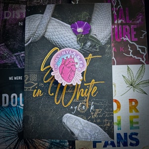 May include: A black and gold sticker with the text "Lost in My Nylakeras" and a pink heart with a white outline. The sticker is on a black background with a white snake.