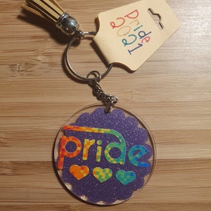 May include: A rainbow pride keychain with a glitter finish and the word "pride" in rainbow colors. The keychain has a small tassel and a silver ring.