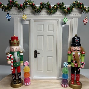May include: Two wooden nutcracker figurines stand on either side of a white door with a Christmas garland above. The nutcrackers are dressed in green and red uniforms with candy canes in their hands. Each nutcracker has a stack of colorful candy on a small stand in front of them.