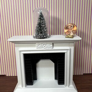 May include: A miniature white fireplace with a black interior and a small Christmas tree inside a glass dome. A penny is placed on the mantel. The background features a striped wallpaper.