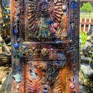 May include: A rectangular wooden panel featuring a heavily textured, multi-colored finish with shades of rust, bronze, and deep blue. The surface is adorned with numerous embedded glass marbles and stones, arranged in two symmetrical sunburst patterns.