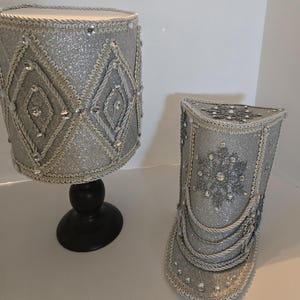 Op de afbeelding: Twee decoratieve zilveren glitter lampenkappen. Eén is cilindrisch en gemonteerd op een zwarte voet. De andere heeft een driehoekige vorm. Beide zijn versierd met zilveren touwranden, strass-steentjes en ruitpatronen.