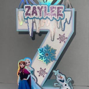 May include: A number 2 birthday cake topper featuring characters from the movie Frozen. The topper has a holographic crown, the name "ZAYLEE" in glittery letters, snowflakes, and images of Anna, Elsa, and Olaf.