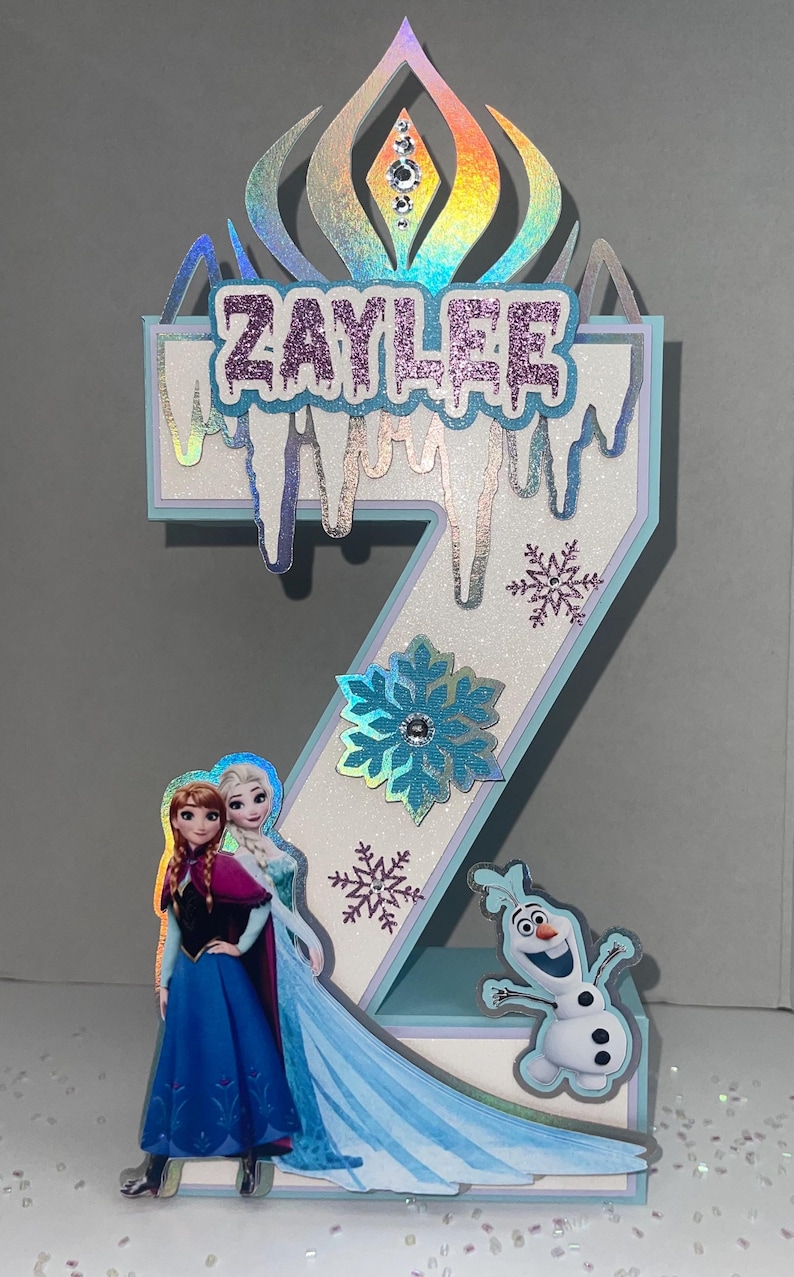 May include: A number 2 birthday cake topper featuring characters from the movie Frozen. The topper has a holographic crown, the name "ZAYLEE" in glittery letters, snowflakes, and images of Anna, Elsa, and Olaf.