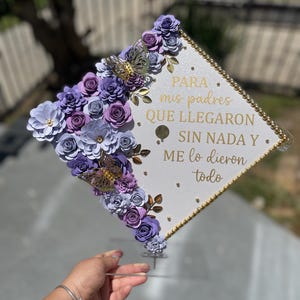 May include: A white graduation cap decorated with purple paper flowers, gold accents, and butterfly embellishments. The cap has gold trim and text in Spanish. A hand holds the cap.