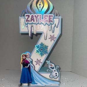 May include: A number two birthday decoration featuring the name "Zaylee" in glittery letters, with images of Elsa, Anna, and Olaf from Frozen. The decoration has a blue and white color scheme with snowflake accents and a holographic crown.