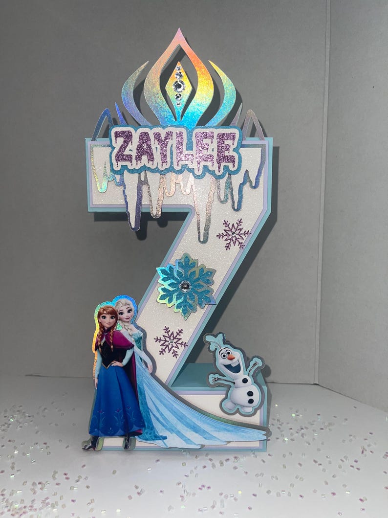 May include: A number two birthday decoration featuring the name "Zaylee" in glittery letters, with images of Elsa, Anna, and Olaf from Frozen. The decoration has a blue and white color scheme with snowflake accents and a holographic crown.