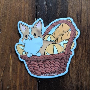 May include: A cartoon illustration of a corgi dog sitting in a wicker basket filled with bread. The corgi is brown and white with a blue and white fluffy tail. The basket is light blue with a brown wicker pattern. The text "MADAMEWITCHART" is at the bottom of the basket.