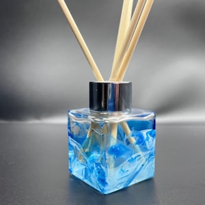 May include: A square glass diffuser bottle with a silver cap and blue liquid. The bottle has a textured, marbled design. Wooden diffuser sticks are inserted into the bottle.
