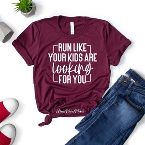 K&ouml;nnte beinhalten: Burgunderfarbenes T-Shirt mit wei&szlig;em Text, der "Lauf, als w&uuml;rden deine Kinder nach dir suchen" lautet. Das Shirt ist auf wei&szlig;em Hintergrund mit Jeans und roten Turnschuhen abgebildet.