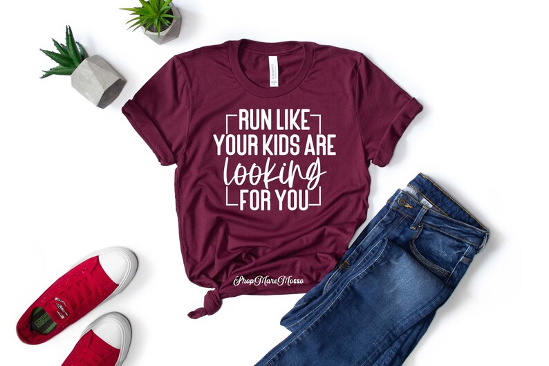 K&ouml;nnte beinhalten: Burgunderfarbenes T-Shirt mit wei&szlig;em Text, der "Lauf, als w&uuml;rden deine Kinder nach dir suchen" lautet. Das Shirt ist auf wei&szlig;em Hintergrund mit Jeans und roten Turnschuhen abgebildet.