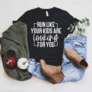 K&ouml;nnte beinhalten: Schwarzes T-Shirt mit wei&szlig;em Text, der "Run like your kids are looking for you" lautet. Das Shirt liegt auf einer wei&szlig;en Oberfl&auml;che mit einem gr&uuml;nen Pullover, einer blauen Jeans und braunen Stiefeln.