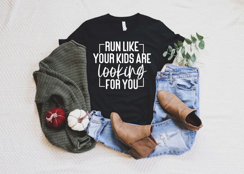 K&ouml;nnte beinhalten: Schwarzes T-Shirt mit wei&szlig;em Text, der "Run like your kids are looking for you" lautet. Das Shirt liegt auf einer wei&szlig;en Oberfl&auml;che mit einem gr&uuml;nen Pullover, einer blauen Jeans und braunen Stiefeln.