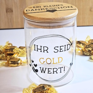 Könnte beinhalten: Ein klares Glas mit einem Bambusdeckel. Auf dem Glas steht in Schwarz und Gold "Ihr seid Gold Wert!" und "Ein kleines Dankeschön". Goldverpackte Süßigkeiten umgeben das Glas.