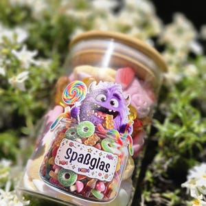 Könnte beinhalten: Ein klares Glas mit bunten Süßigkeiten, darunter Gummibärchen, Lollis und ringförmige Süßigkeiten. Ein Aufkleber auf dem Glas zeigt ein lila Monster und das Wort "Spaßglas". Das Glas hat einen Holzdeckel und steht vor grünem Laub.