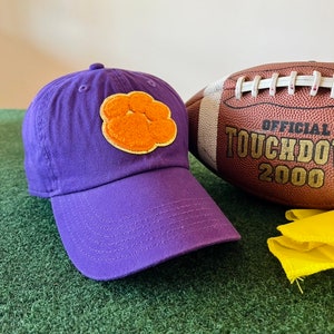 May include: Purple baseball cap with an orange paw print embroidered on the front. The cap is sitting on a green surface next to a brown football with the text "OFFICIAL TOUCHDOWN 2000".