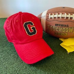 May include: A red baseball cap with a black and gold letter "G" patch is next to a brown football with white laces. The football has the text "OFFICIAL TOUCHDOWN 2000". The items are on a green surface.
