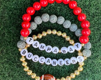 Puede incluir: Tres pulseras superpuestas con cuentas rojas, grises y doradas. La pulsera del medio tiene cuentas blancas con las palabras "Tampa Bay Buccaneers" escritas. La pulsera de abajo tiene un dije de fútbol.
