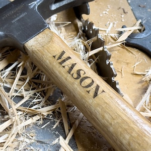 May include: A wooden-handled hammer with a black metal head. The handle is engraved with the name "MASON". The hammer is lying on a surface covered in wood shavings.