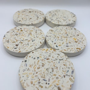 May include: Five round coasters with a white base, speckled with a mosaic of seashells in shades of beige, brown, and gold. The coasters have a smooth, slightly raised edge. These decorative coasters are ideal for protecting surfaces.