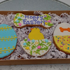 May include: Four Easter-themed dog biscuits. One is a bone-shaped biscuit with the words "Happy Easter!" in purple icing. Another is a round biscuit with a yellow chick in a nest of green and pink sprinkles. The third is a round biscuit with a blue and yellow egg design. The last is a round biscuit with a white base and yellow and pink sprinkles.