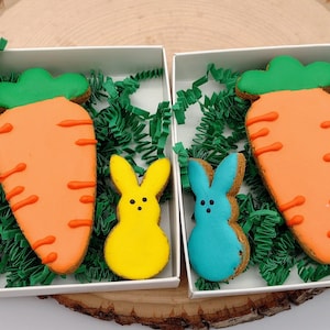 May include: A set of Easter-themed dog treats in a white box. The treats are shaped like carrots and bunnies. The bunnies are pink, yellow, blue, and purple. The carrot is orange with green leaves.