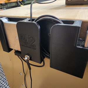 May include: A black 3D printed cable management system with a silver metal clamp. The system has two compartments, one for holding a coiled cable and the other for holding a small tool. The system is mounted on a wooden surface. The system has the text "42 Workshop" printed on it.