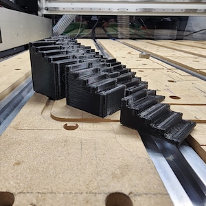 May include: A black plastic 3D printed step-up block with multiple levels. The block is made of several stacked pieces, each with a slightly different height. The block is sitting on a wooden surface.