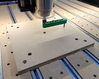 May include: A close-up of a CNC machine with a green clamp holding a beige wood panel. The panel has a series of holes and the text "150 WO 116955 07/12/2018 STD2" printed on it.