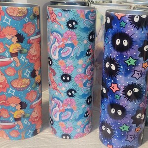 May include: Three stainless steel tumblers with colorful anime-style designs. One features a blue background with characters and food, another has a light blue background with a dragon and flowers, and the last has a dark blue background with star-shaped figures.