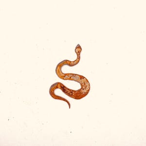 May include: A rusty metal snake sculpture with a textured surface. The snake is coiled in a stylized S-shape, with a small, pointed head and a tapering tail. The sculpture is set against a plain, off-white background.