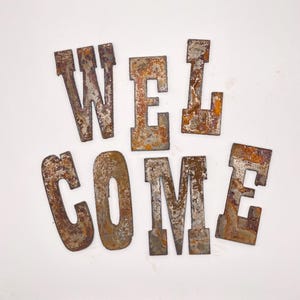 May include: Rusty metal letters spelling "WELCOME" arranged on a white surface. The letters have a distressed, aged appearance with shades of brown, orange, and gray, suggesting an industrial or vintage aesthetic. The letters are in a bold, blocky font.